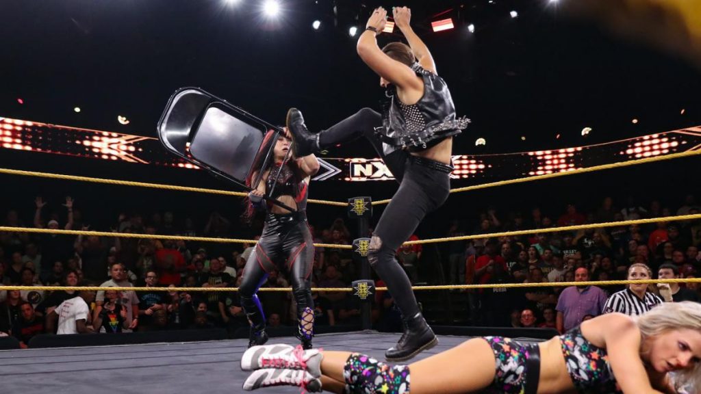 Rhea Ripley kicks a chair out of Io Shirai's hand, with Candice LeRae in the foreground