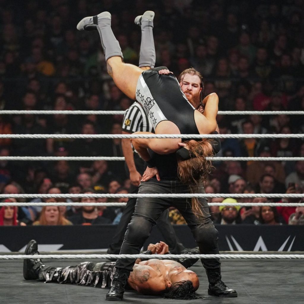 Killian Dain holds Pete Dunne upside down with Damia Priest laying at his feet.