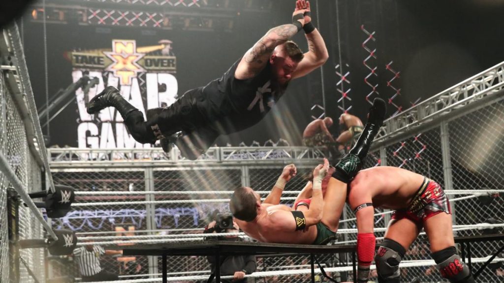 Kevin Owens splashes Kyle O'Reilly through a table to get him to let go of Dominik Dijakovic. Adam COle and Tommaso Ciamp on top of the cage in the background