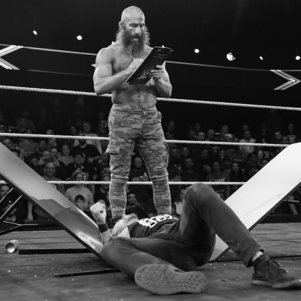 Tommaso Ciampa stads over Adam Cole after putting him through a table