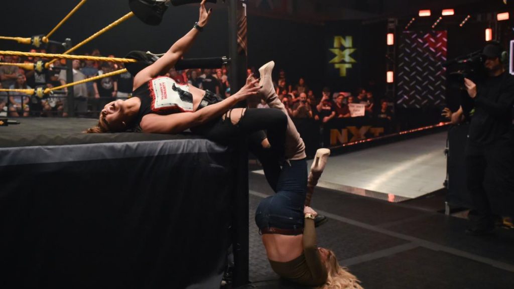 Charlotte Flair twists Rhea Ripley's legs aroud the post and hangs off them