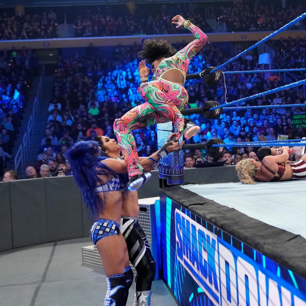 Naomi comes over the top rope onto Sasha Banks