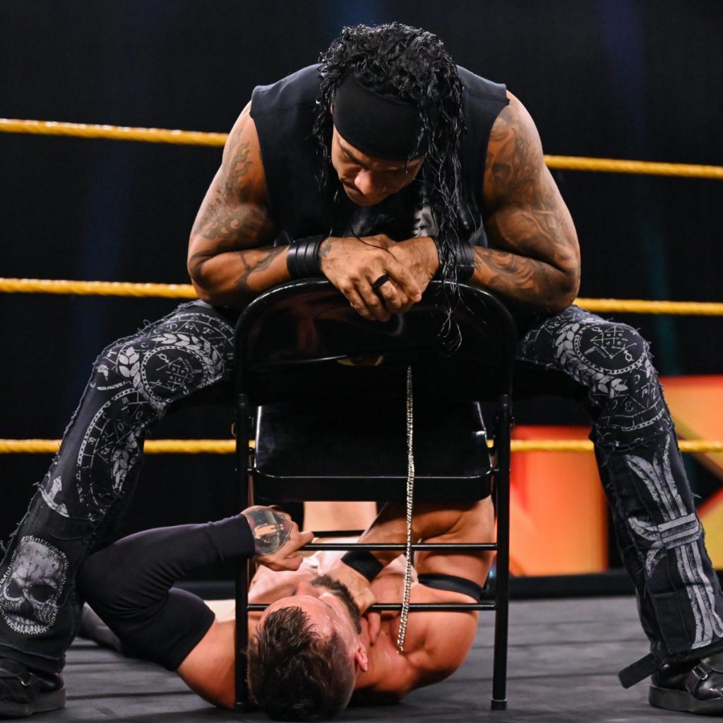 Damian Priest pins Finn Balor under a chair by sitting on it.
