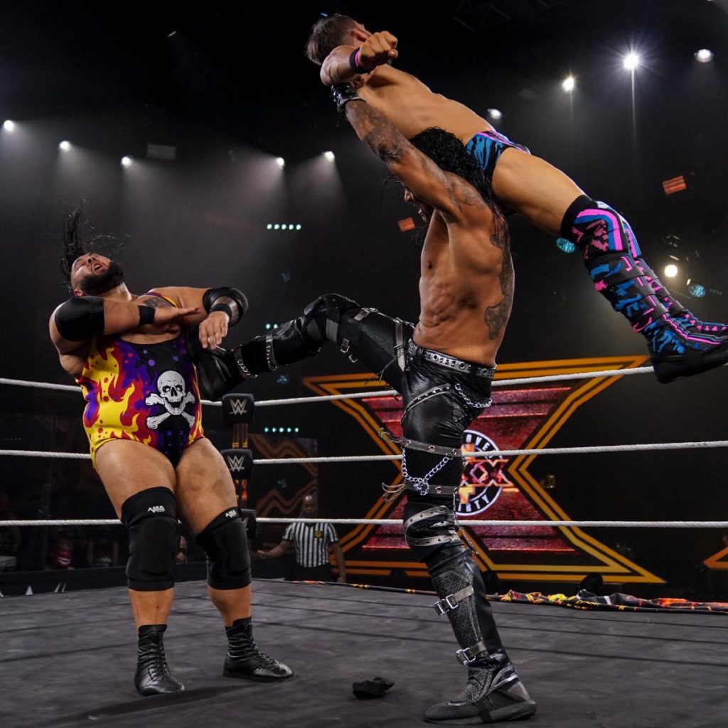 Damian Priest kicks Bronson Reed while holding Johnny Gargano over his head