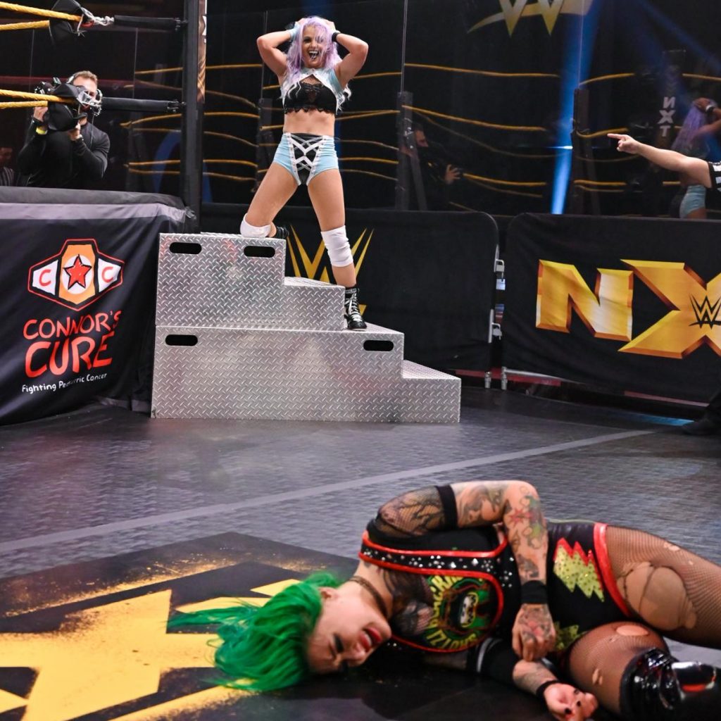 Candice LeRae looks at Shotzi Blackheart laying on the floor, from the steps