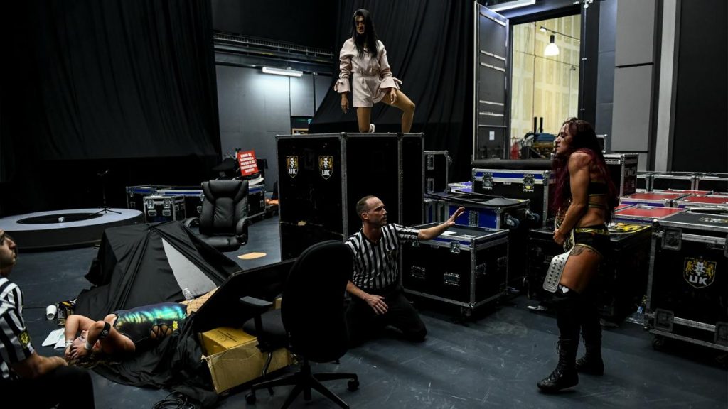 Jinny on top of the packing crate looking at Kay Lee Ray holding the title and Piper Niven laid in the wreckage of a table