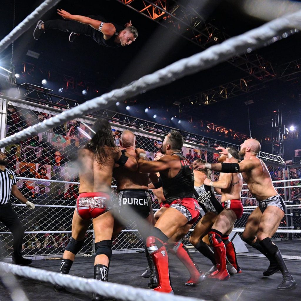 Pat McAfee dives off the top of the cage onto the rest of the teams