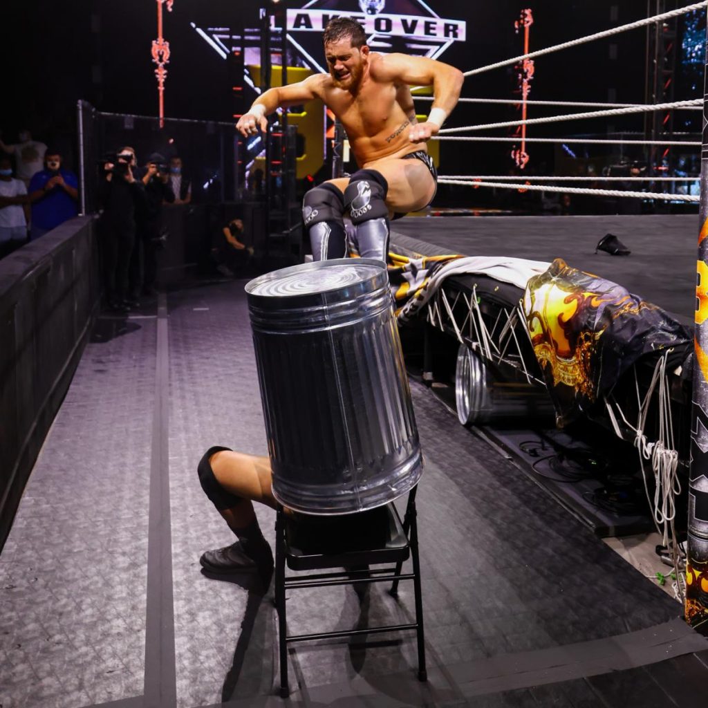 Adam Cole sits on a chair with a trash can on his head as Kyle O'Reilly is about to dropkick him