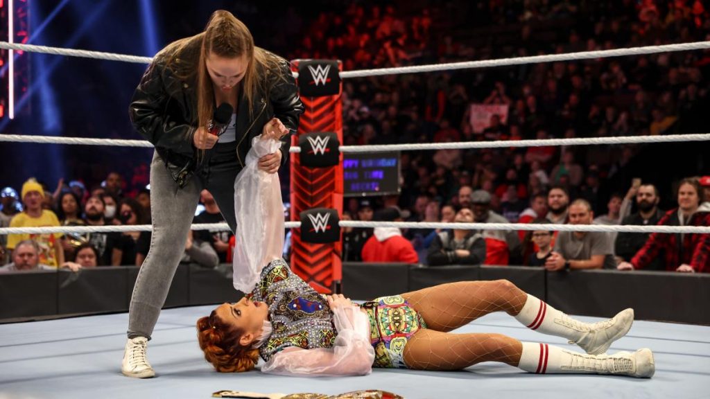 Ronda Rousey stands over Becky Lynch with Lynch's arm locked in a hold